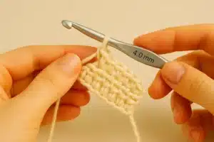 Hands holding a 4.0 mm crochet hook and a small half double crochet swatch in cream yarn