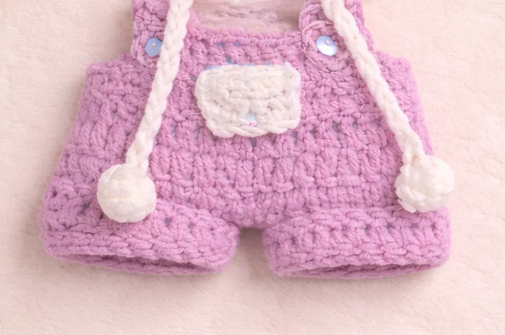 Labubu clothes crochet pattern – purple overalls with white pocket, straps, buttons, and pom-pom ties (flat lay)