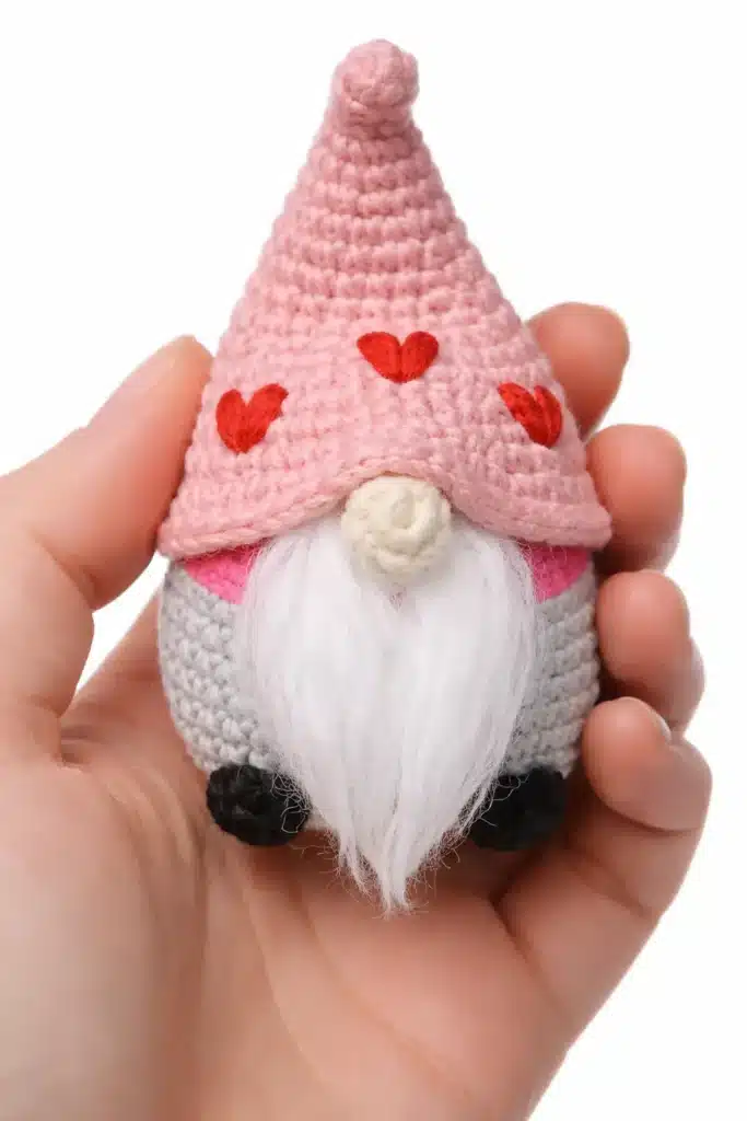 Valentine gnome crochet pattern mini gnome held in hand, with pink hat hearts, beige nose, fluffy beard, and black feet.