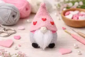 Valentine gnome crochet pattern finished mini gnome with pink heart hat, gray body, and fluffy white beard on a craft table.