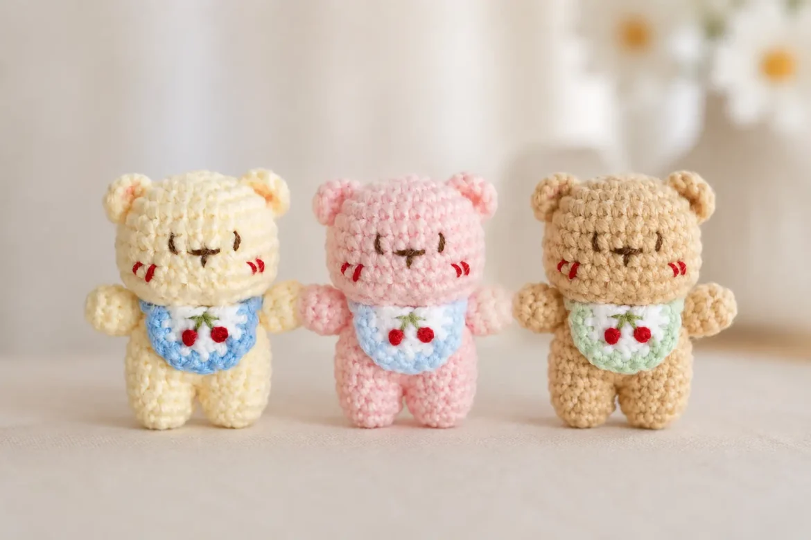 Three Little Bear with Cherry Bib Crochet Pattern color variations in cream, pink, and beige, each wearing a small cherry bib with embroidered red cherries.