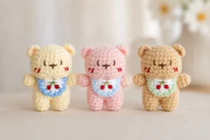 Little Bear with Cherry Bib Crochet Pattern Three Little Bear with Cherry Bib Crochet Pattern color variations in cream, pink, and beige, each wearing a small cherry bib with embroidered red cherries.