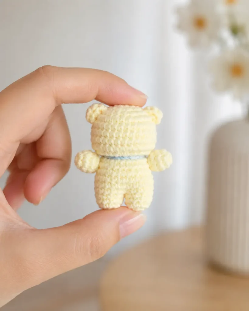 Back view of the Little Bear with Cherry Bib Crochet Pattern showing the tiny cream crochet bear held by hand with rounded ears, small arms, legs, and a blue neck tie.