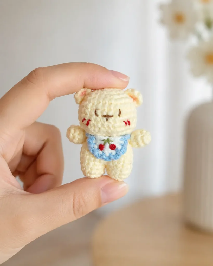 Little Bear with Cherry Bib Crochet Pattern showing a tiny cream crochet bear held by hand with a blue bib, red cherry embroidery, stitched face, and rosy cheeks.